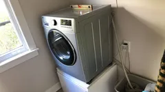 Moderní pračka v rohu prádelny poblíž okna, elektro zásuvka na zdi.